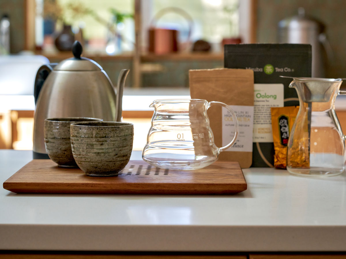 Tea kettle, cups, and glass pitchers set up on a countertop with different bags of tea behind them.