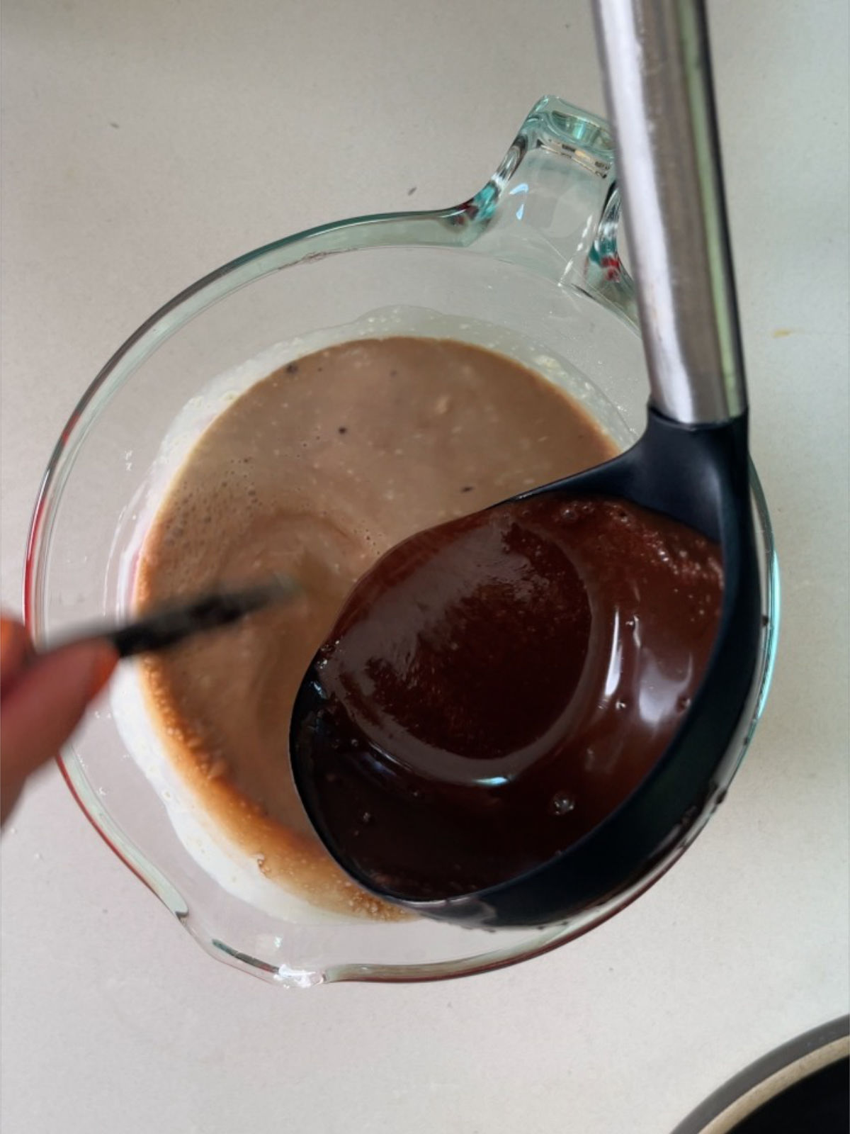 Black and metal ladle pouring chocolate liquid into a glass measuring cup creating a lighter brown liquid in the cup.