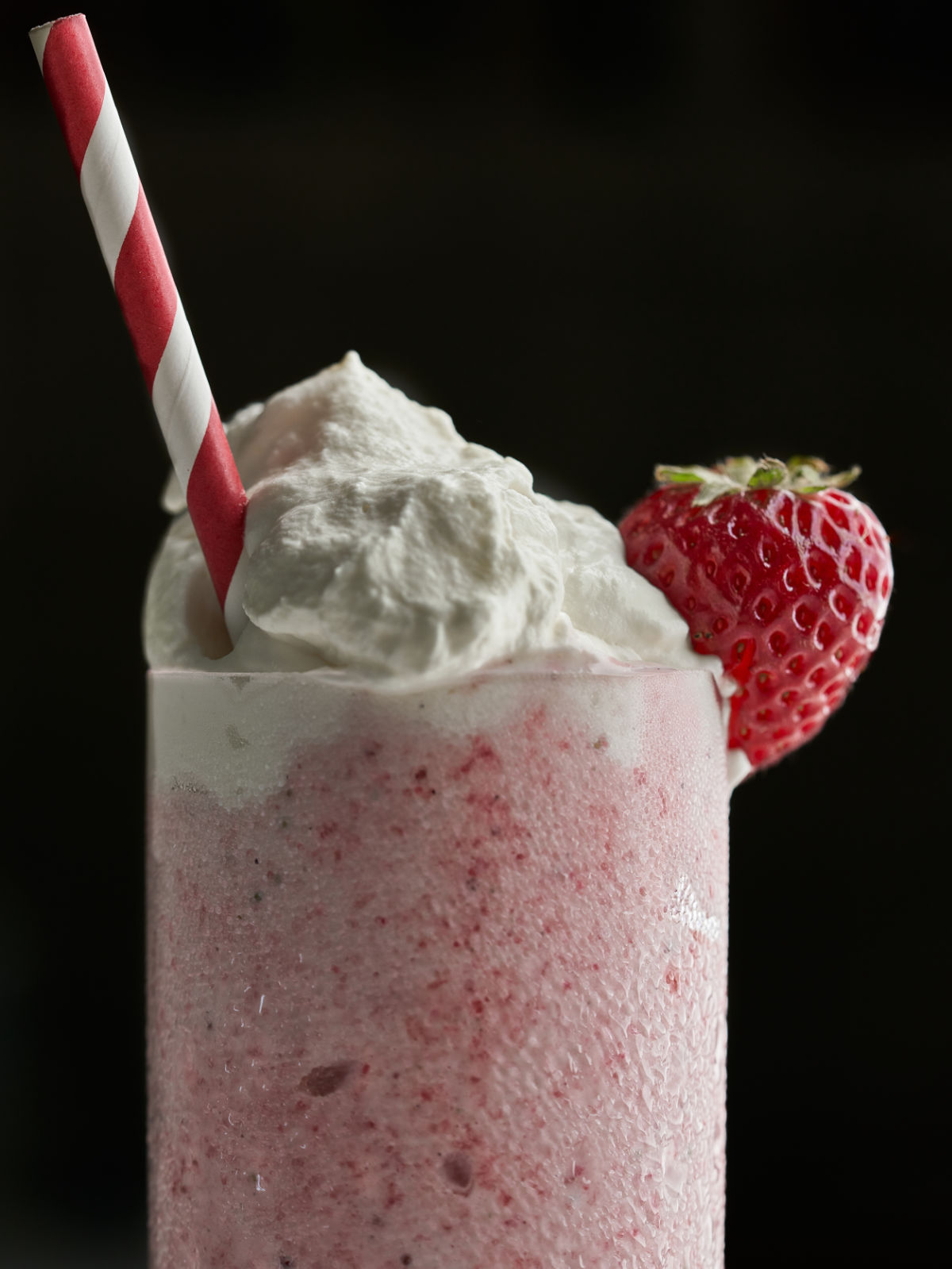 Pink milkshake with a whipped cream topping, strawberry, and red and white striped straw.