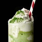 Green milkshake with a whipped cream topping sprinkled with green powder, and red and white striped straw.