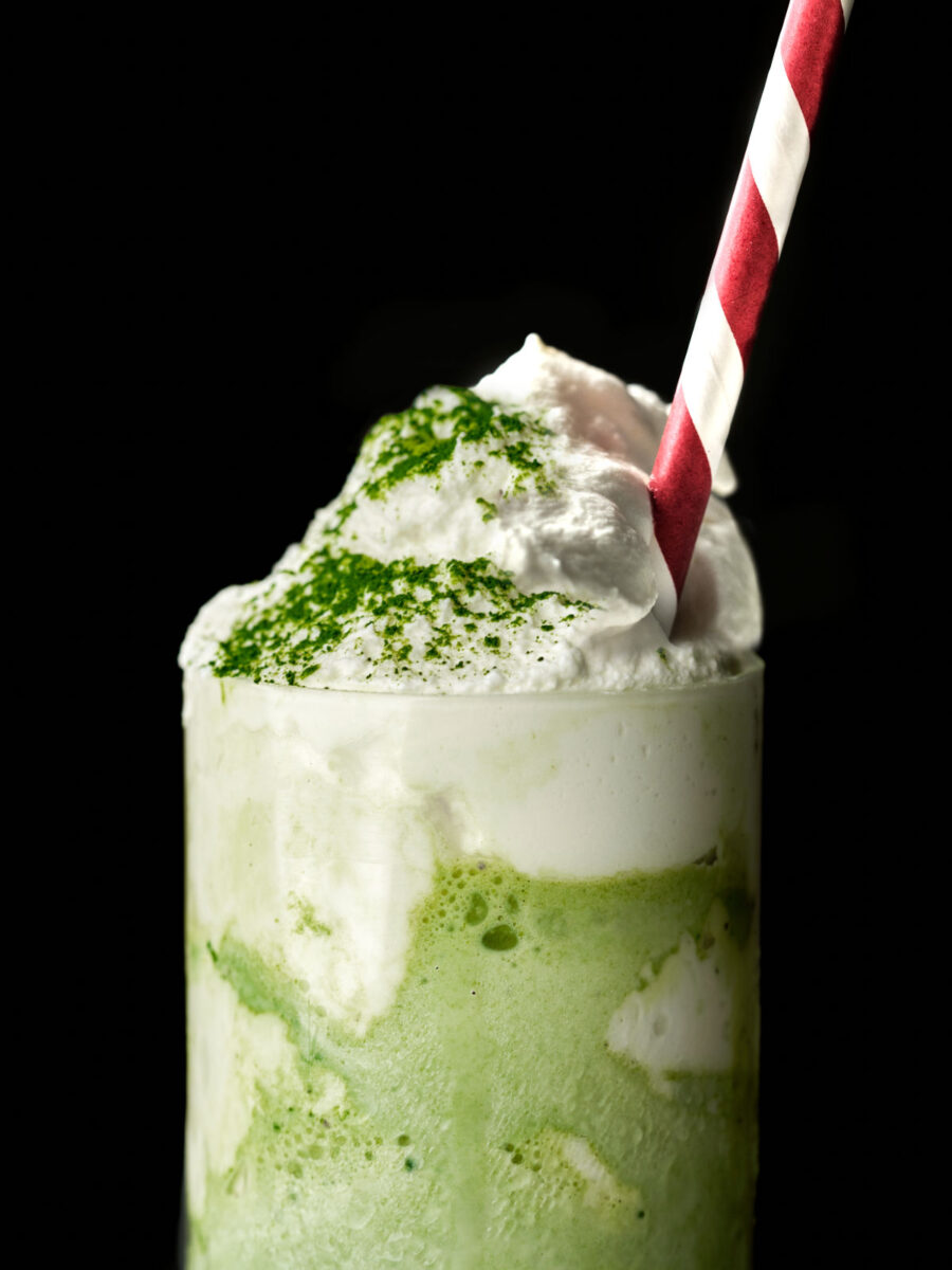 Green milkshake with a whipped cream topping sprinkled with green powder, and red and white striped straw.