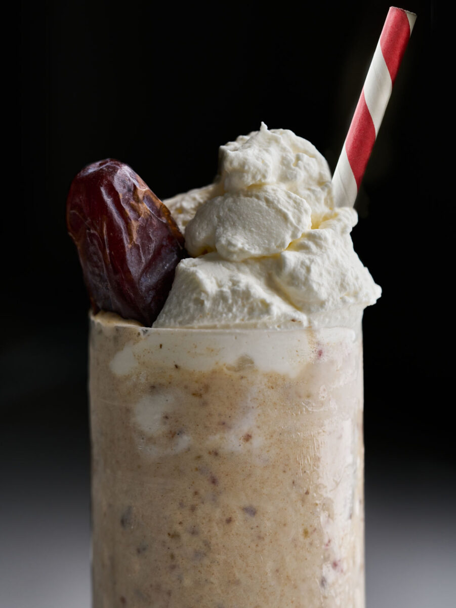Light brown milkshake with a whipped cream topping, date garnish, and red and white striped straw.