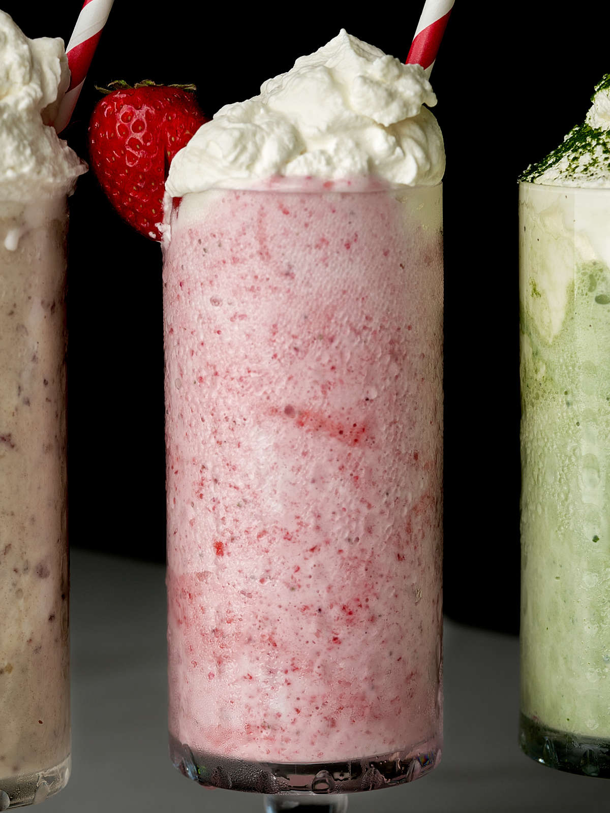 Pink milkshake in between and green milkshake and a light brown milkshake, all with red and white striped straws.