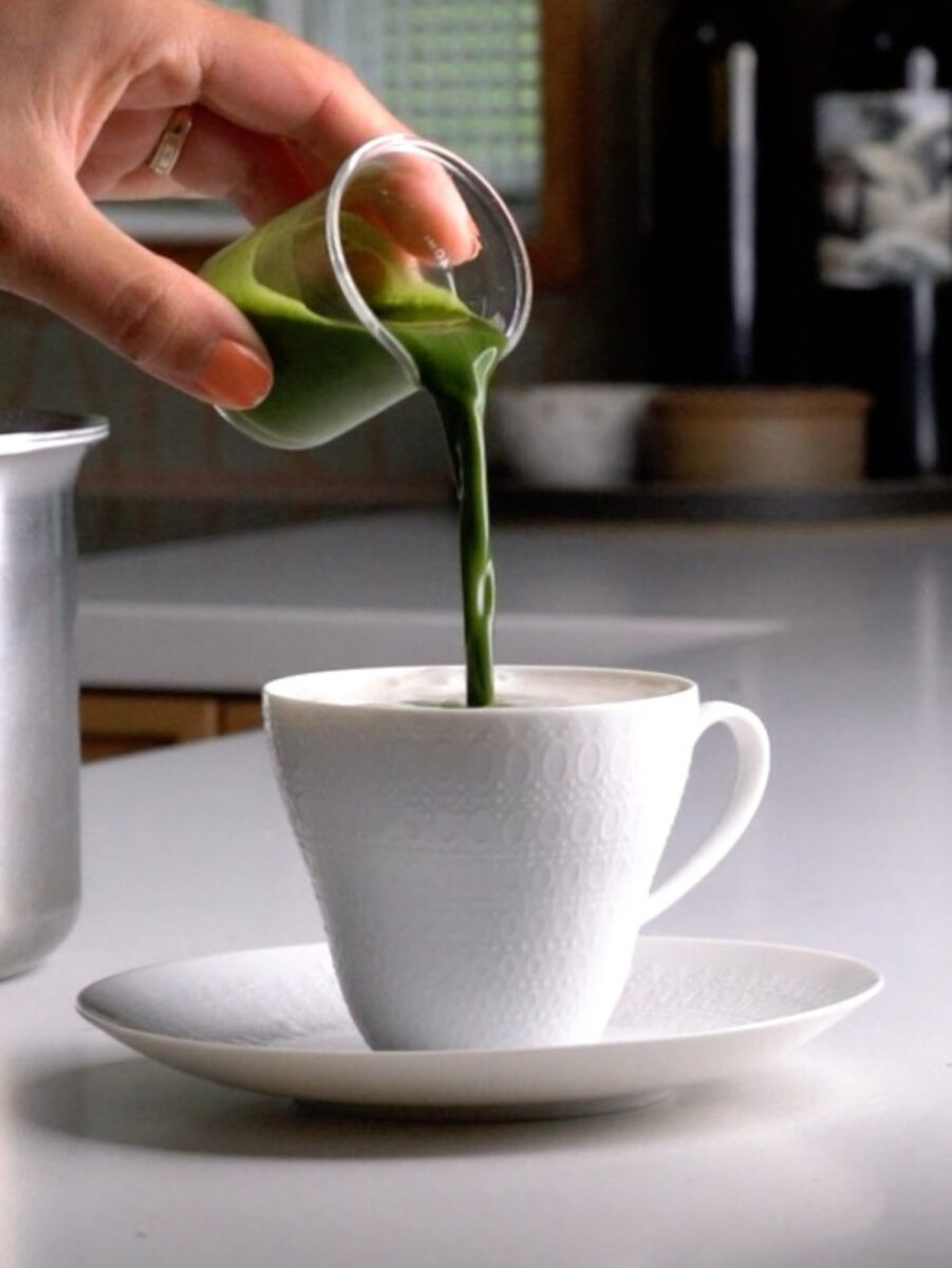 Pouring a shot of matcha into a porcelain tea cup resting on a saucer.