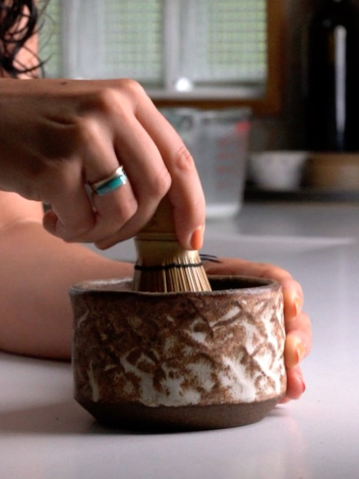 Hand with a turquoise ring whisking matcha in a brown ceramic bowl with a matcha whisk.