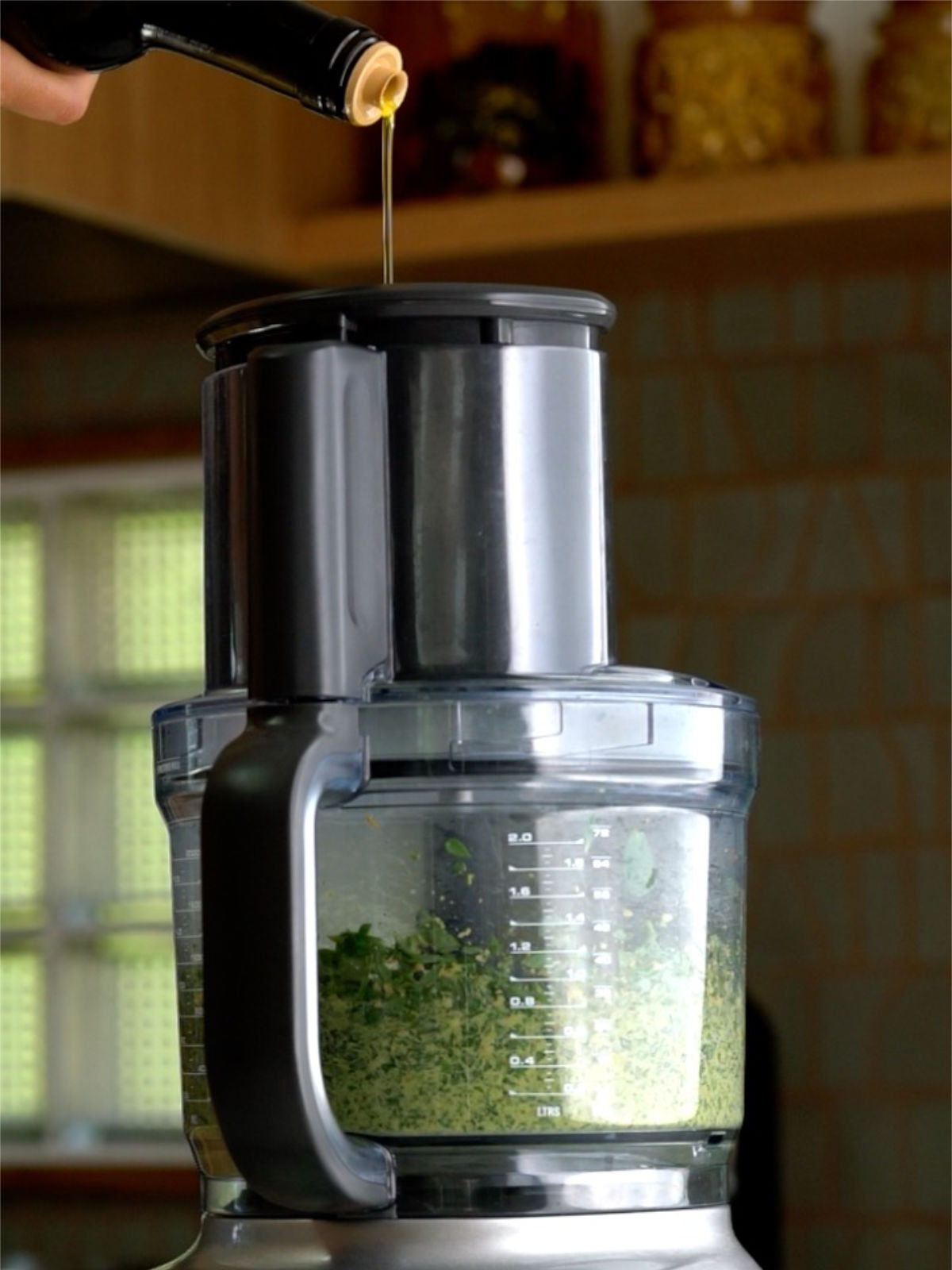 Pouring olive oil into the top of a food processor with green pesto in the bowl.