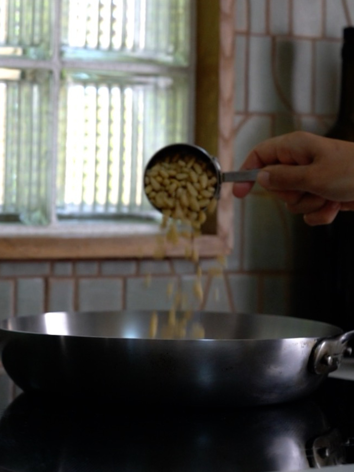 Pouring pin nuts from a metal measuring cup into a metal pan on a cooktop.