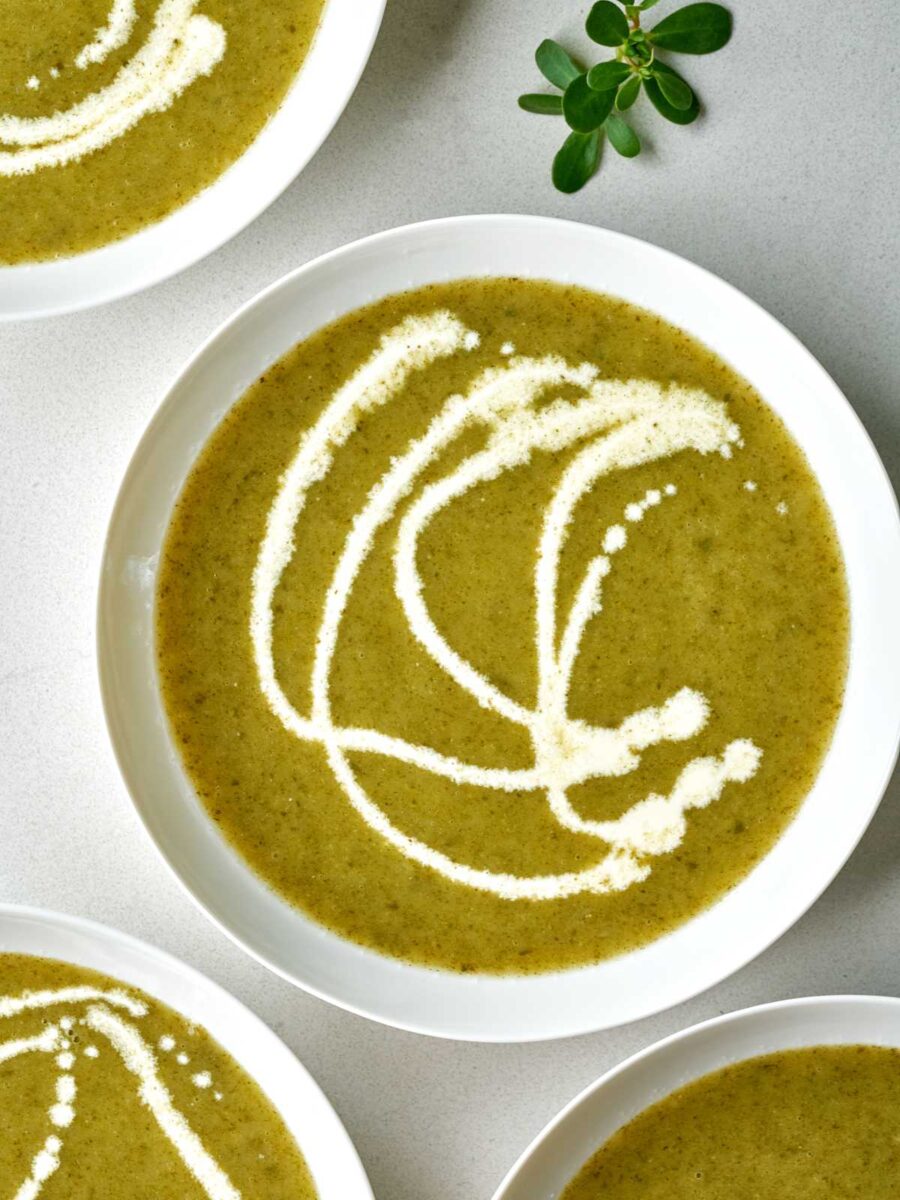 Green pureed soup in a white bowl with white cream drizzle next to a sprig of purslane.