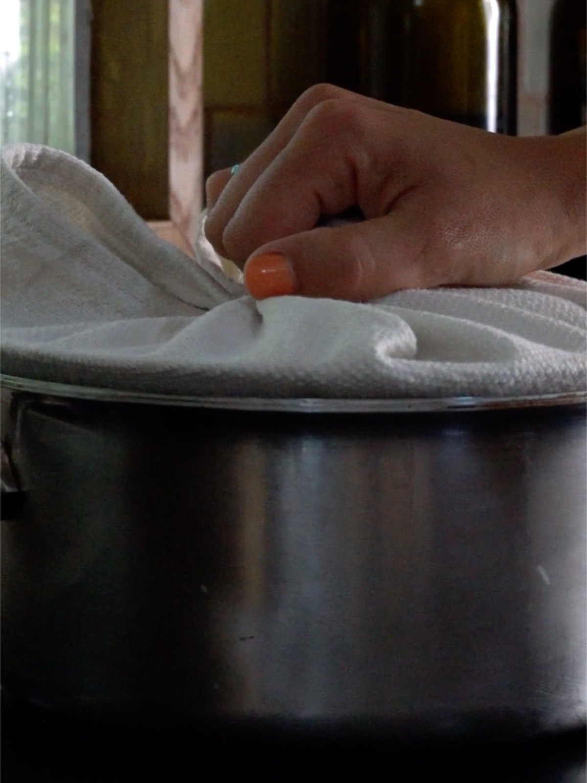 Hand with orange nail polish holding the lid of a metal pot wrapped in a white towel on a metal pot.