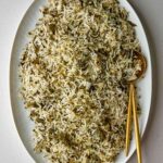 Rice mixed with herbs on an oval white platter with gold serving utensils.