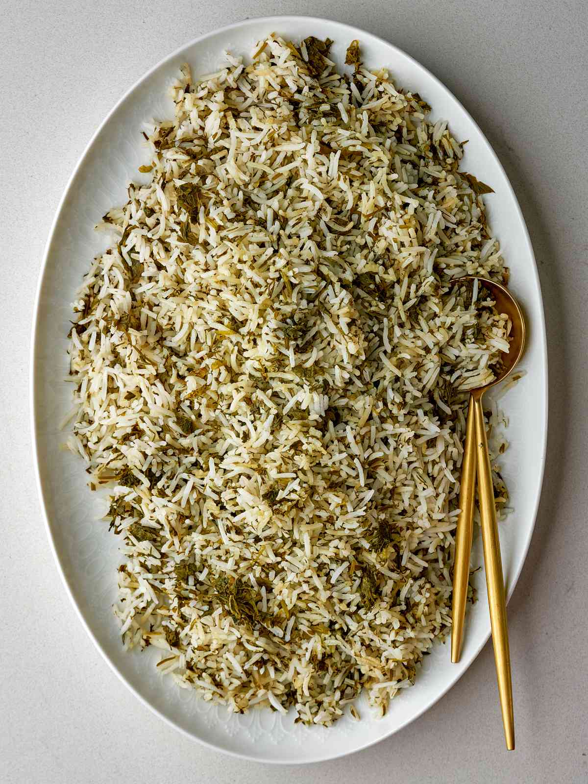 Rice mixed with herbs on an oval white platter with gold serving utensils.