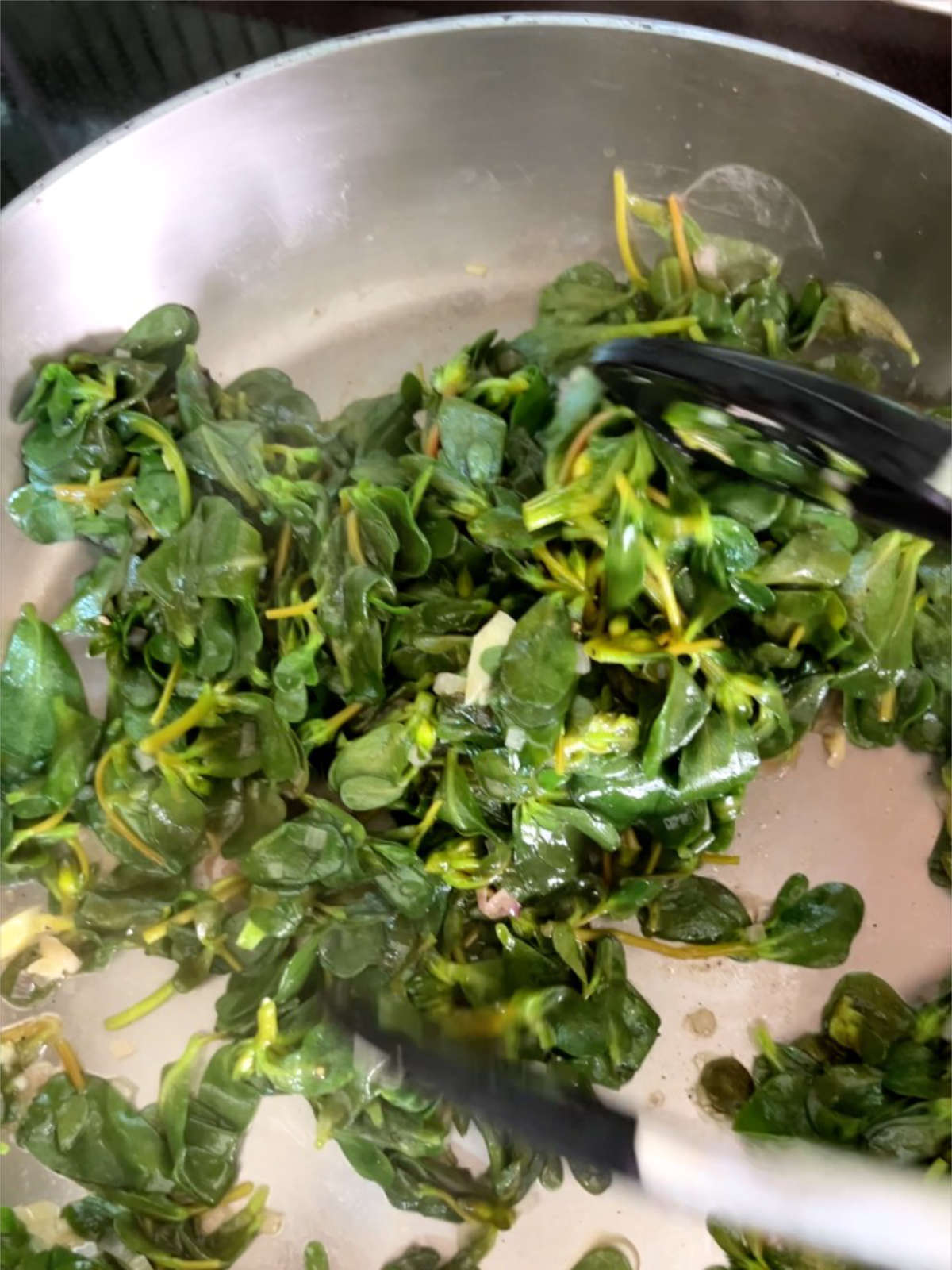 Wilted greens in a metal pan with black and metal tongs.