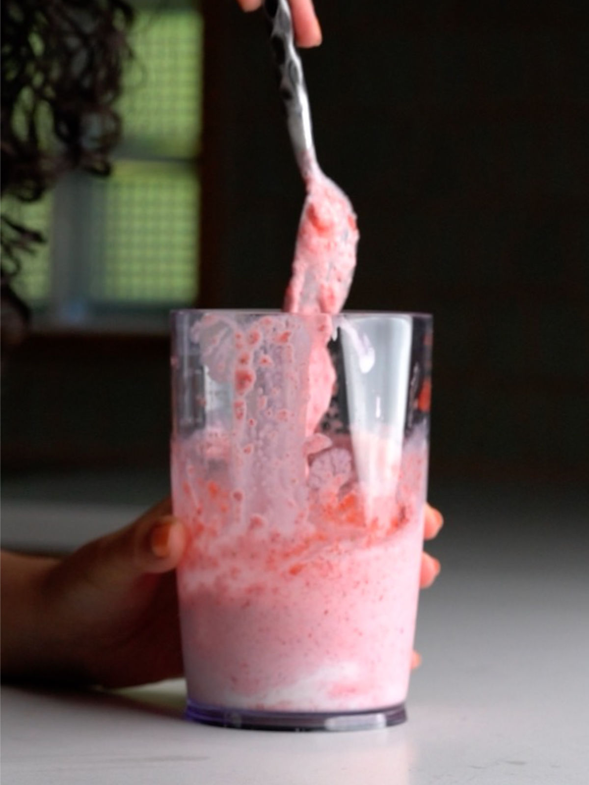 Pink strawberry milkshake in a clear immersion blender container being stirred with a spoon.