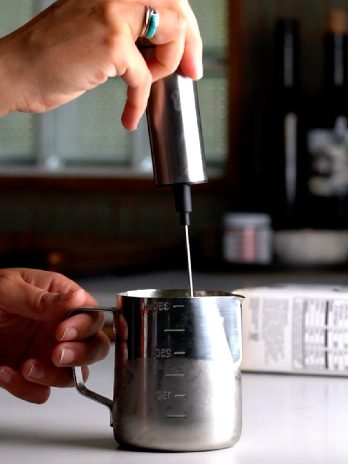 A handheld frother frothing liquid in a metal measuring cup.