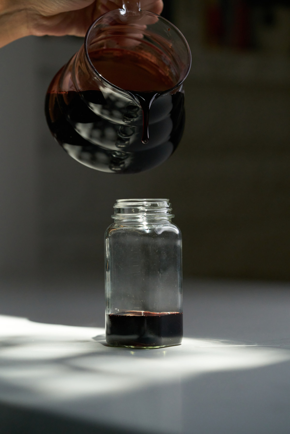 Pouring burgundy syrup into a small glass jar from a glass pitcher.