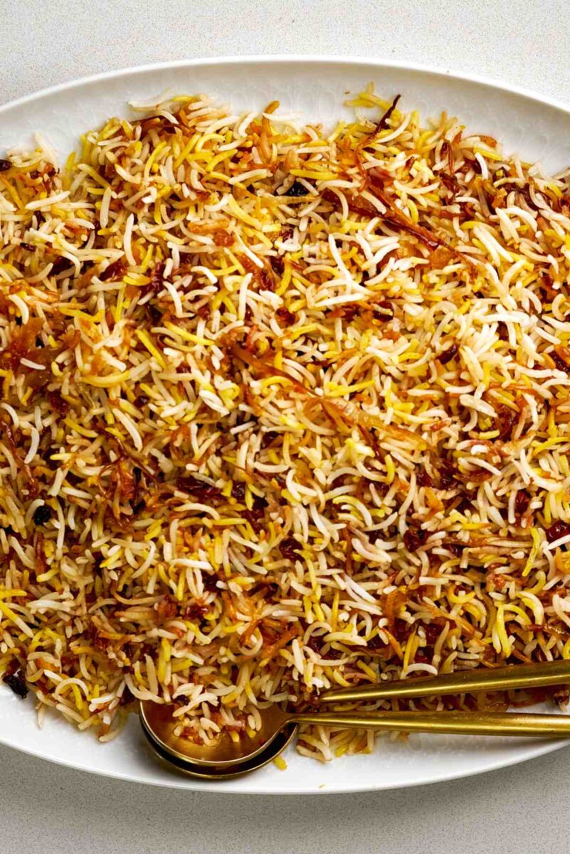 Rice pilaf on a white oval platter with gold serving utensils.