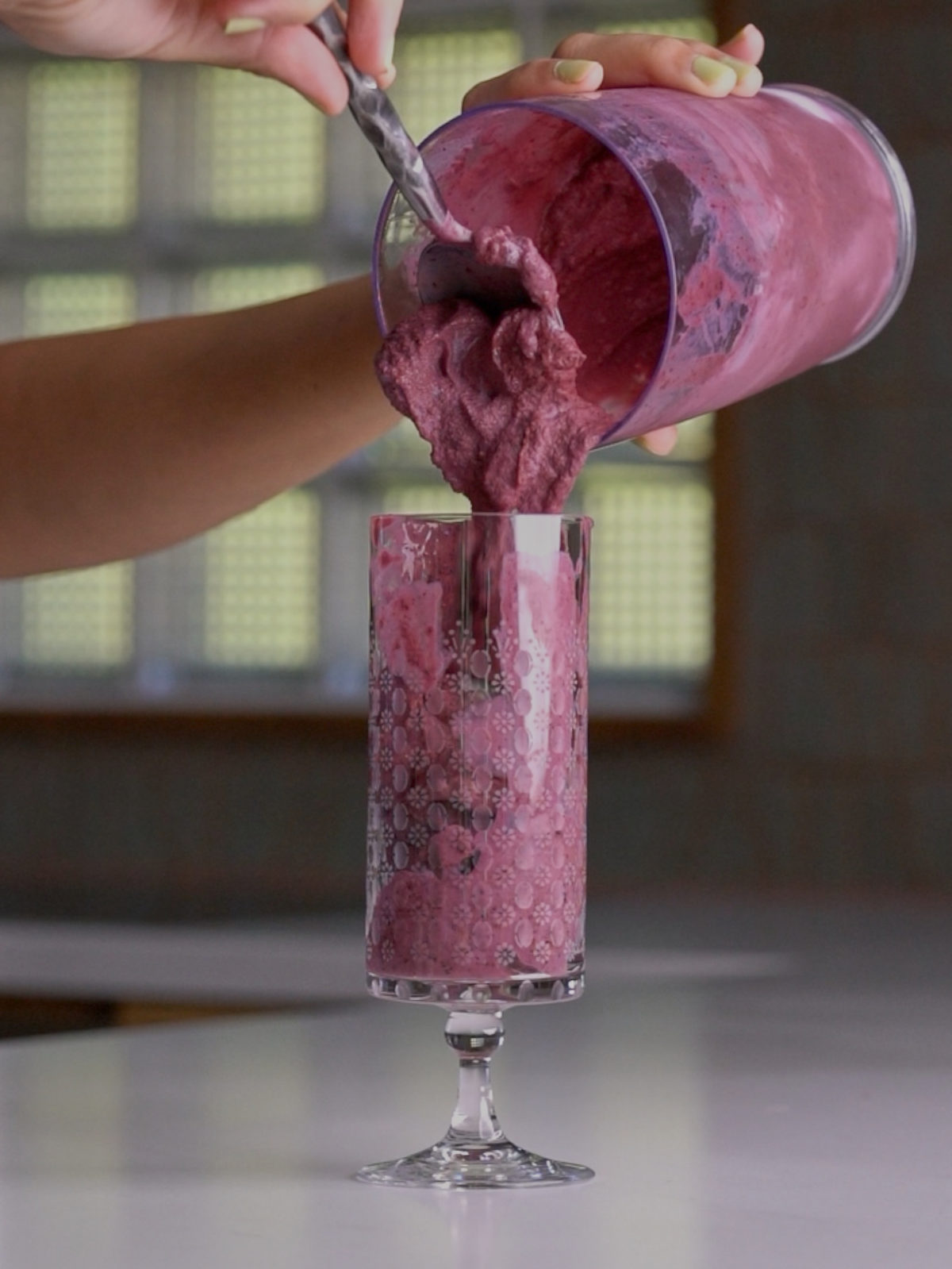 Pouring blackberry milkshake into a clear pedestal glass with a spoon.