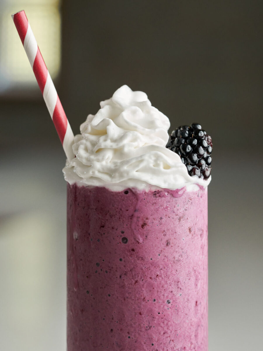 Purple milkshake topped with whipped cream, a blackberry, and a striped straw.