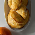 Pile of half-moon shaped hand pies on a white plate next to a small orange pumpkin.