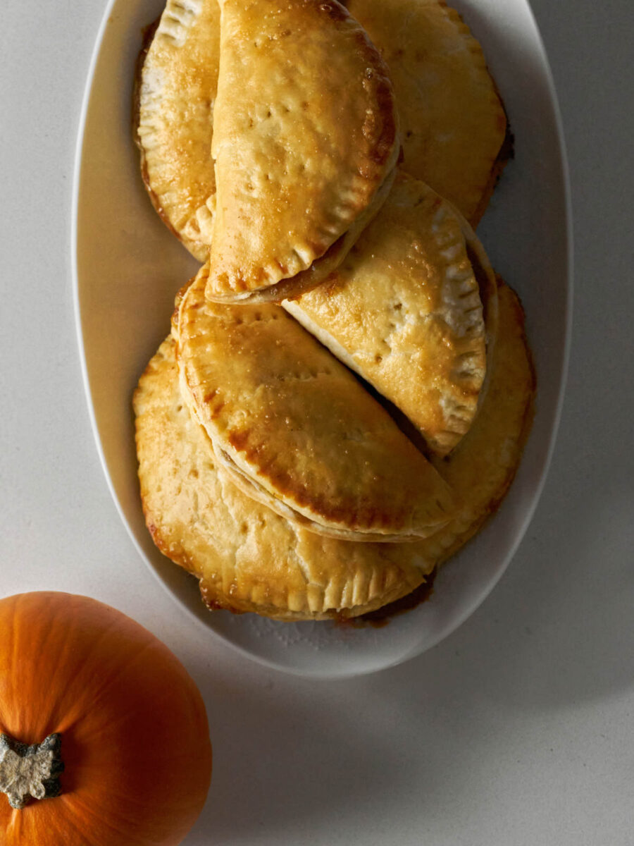 Pile of half-moon shaped hand pies on a white plate next to a small orange pumpkin.
