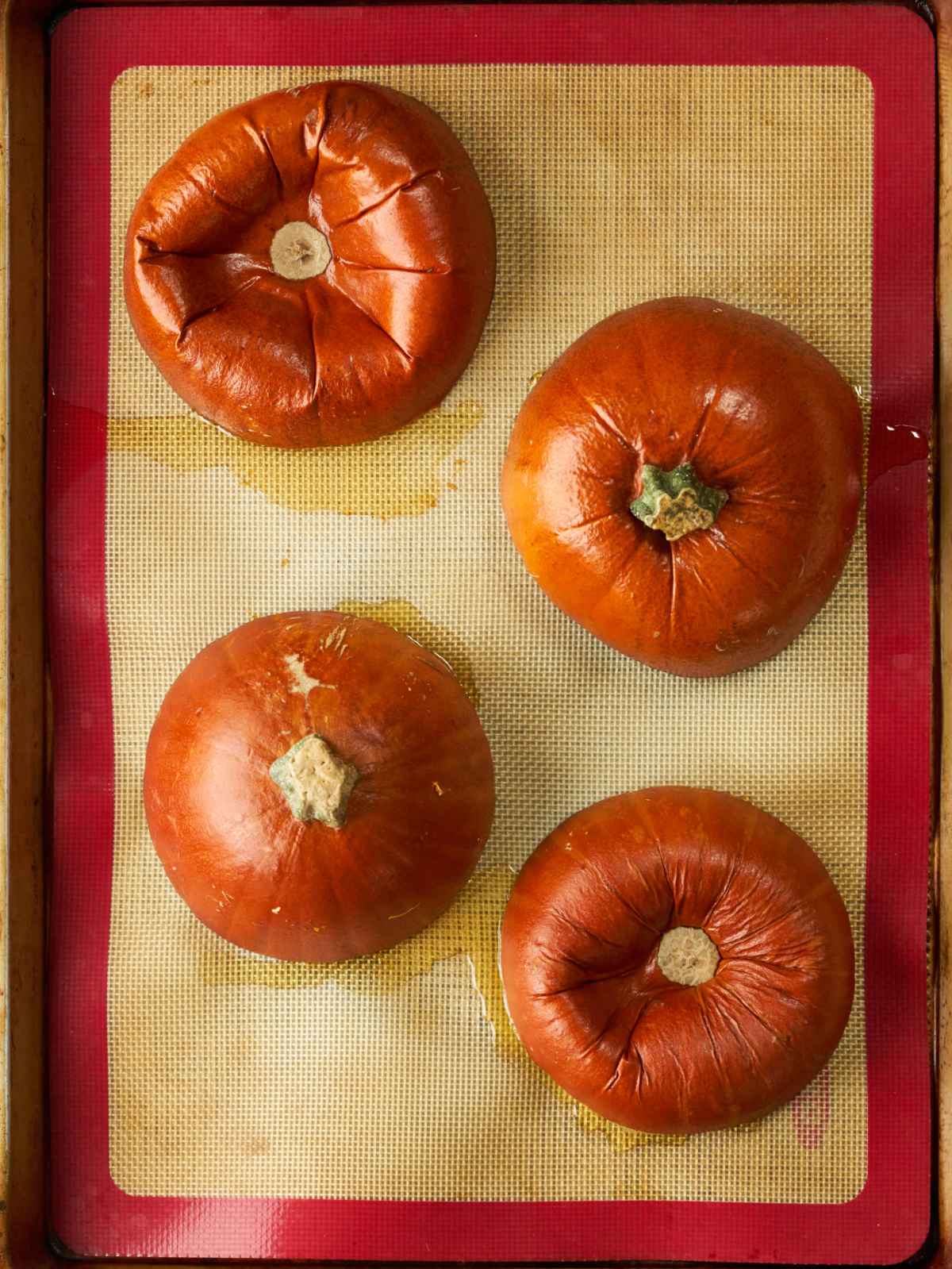 Four baked pumpkin halves cut side down on a silicone baking sheet.