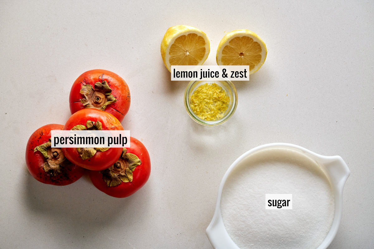 A stack of persimmons next to a sliced lemon with zest and a bowl of sugar.