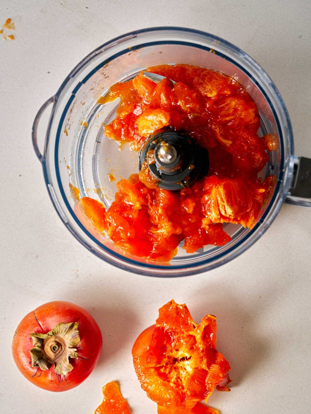 Food processor bowl filled with persimmon pulp next to a persimmon and another peeled persimmon.