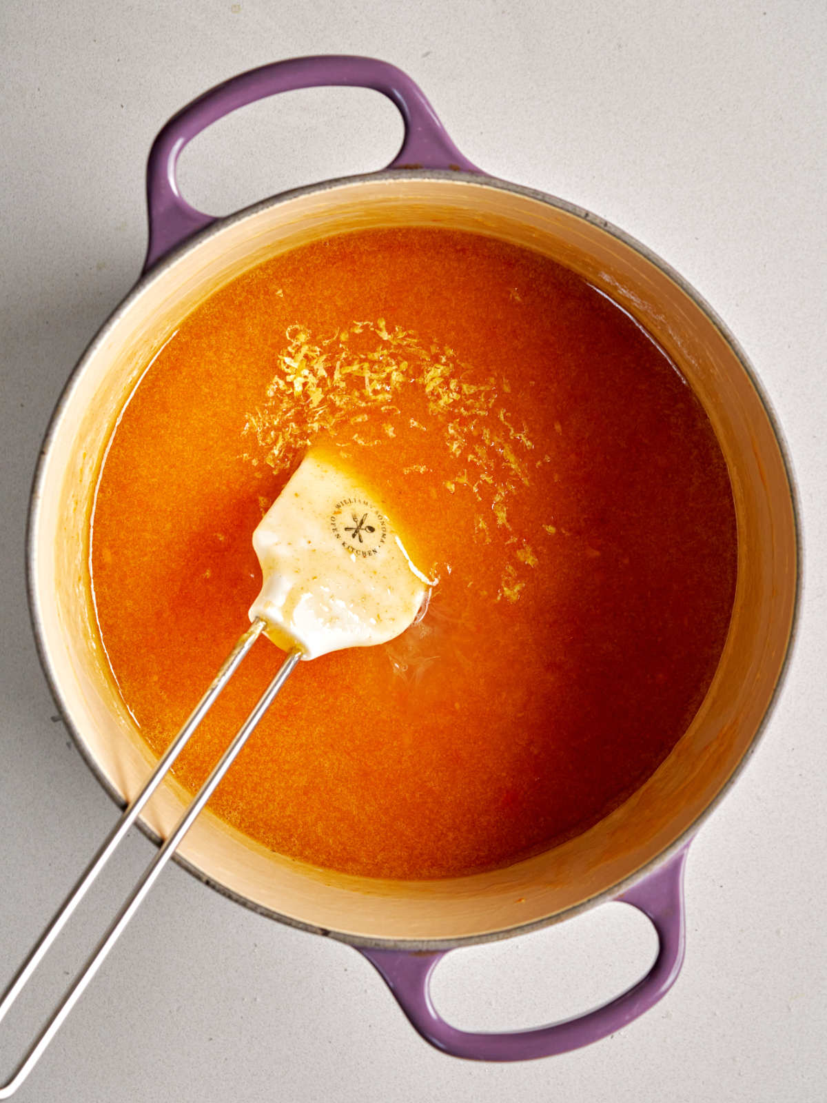 Orange puree in a purple pot with a sprinkle of yellow lemon zest and a white rubber spatula.