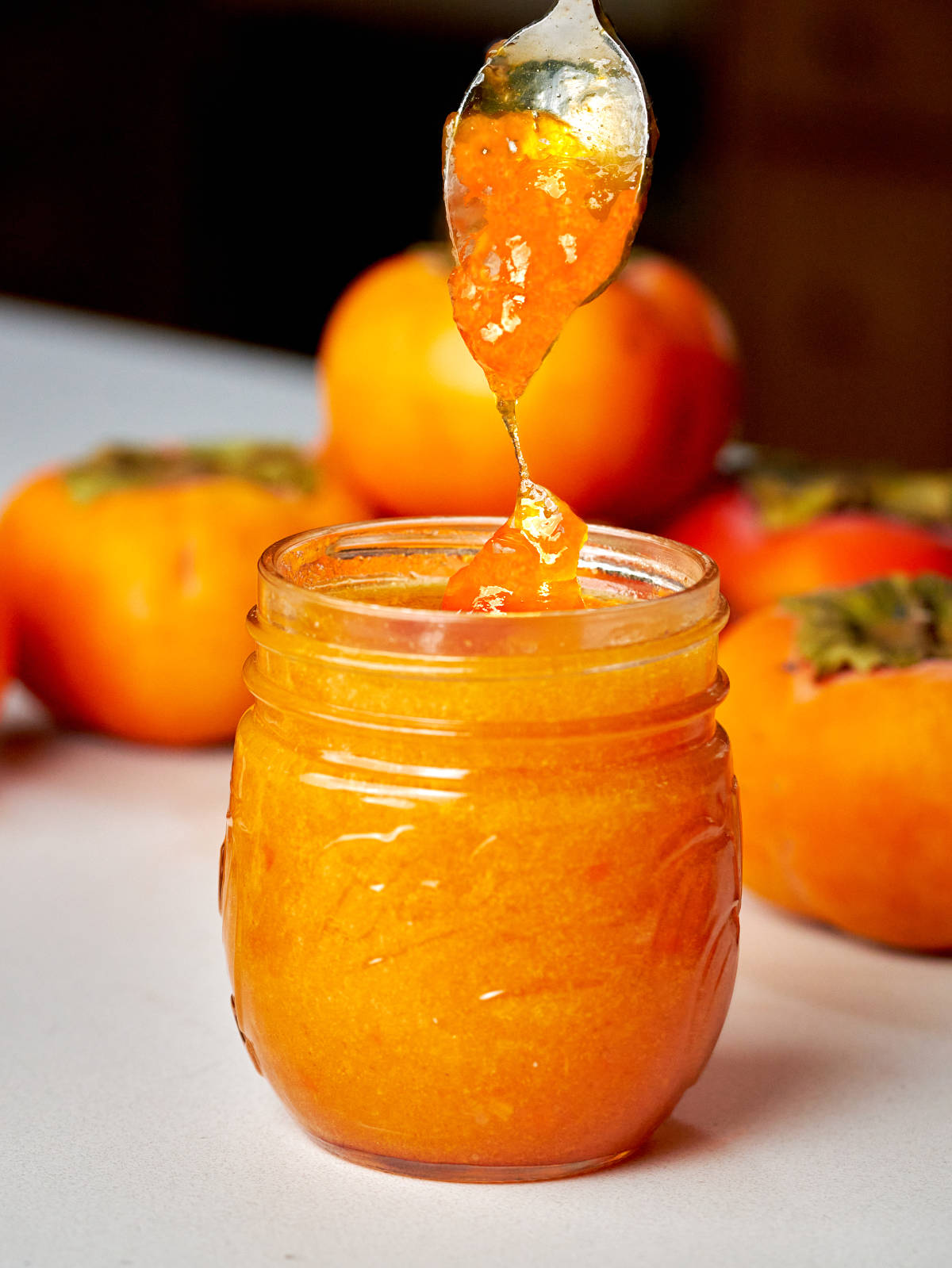 Spoon of orange jam over a small jar of jam in front of a stack of persimmons.