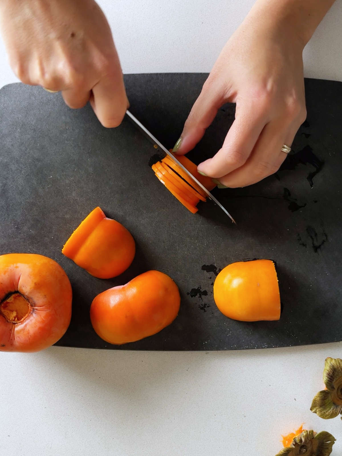 Thinly slicing a Fuyu persimmon on a black cutting board next to persimmon stems.