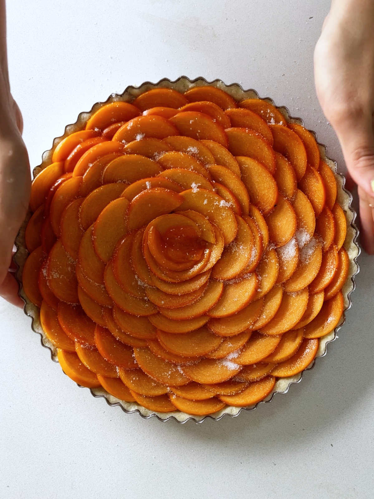 Two hands holding a tart filled with orange fruit arranged in a rose pattern.