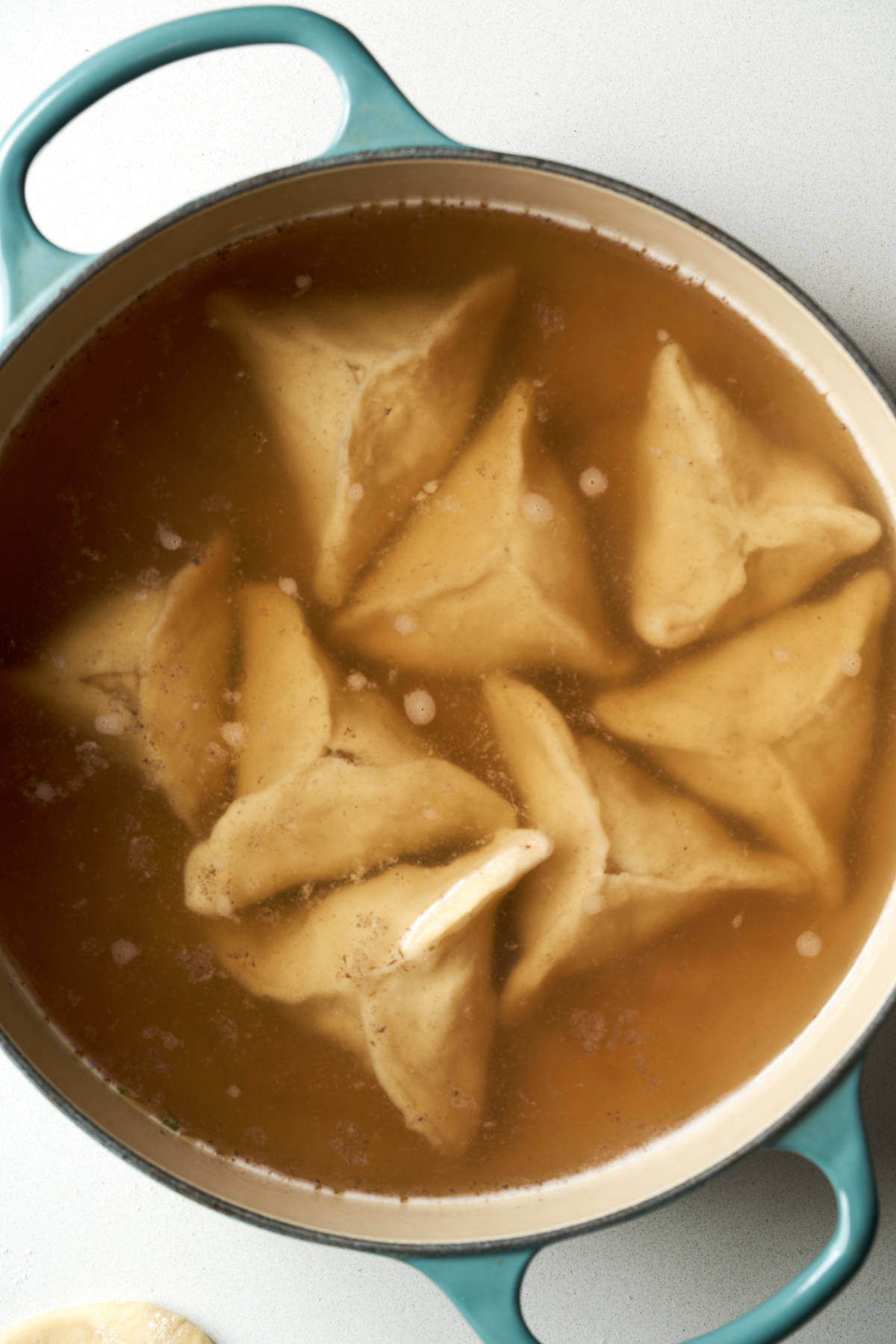 Triangular kreplach dumplings in chicken broth in a teal cast iron pot.