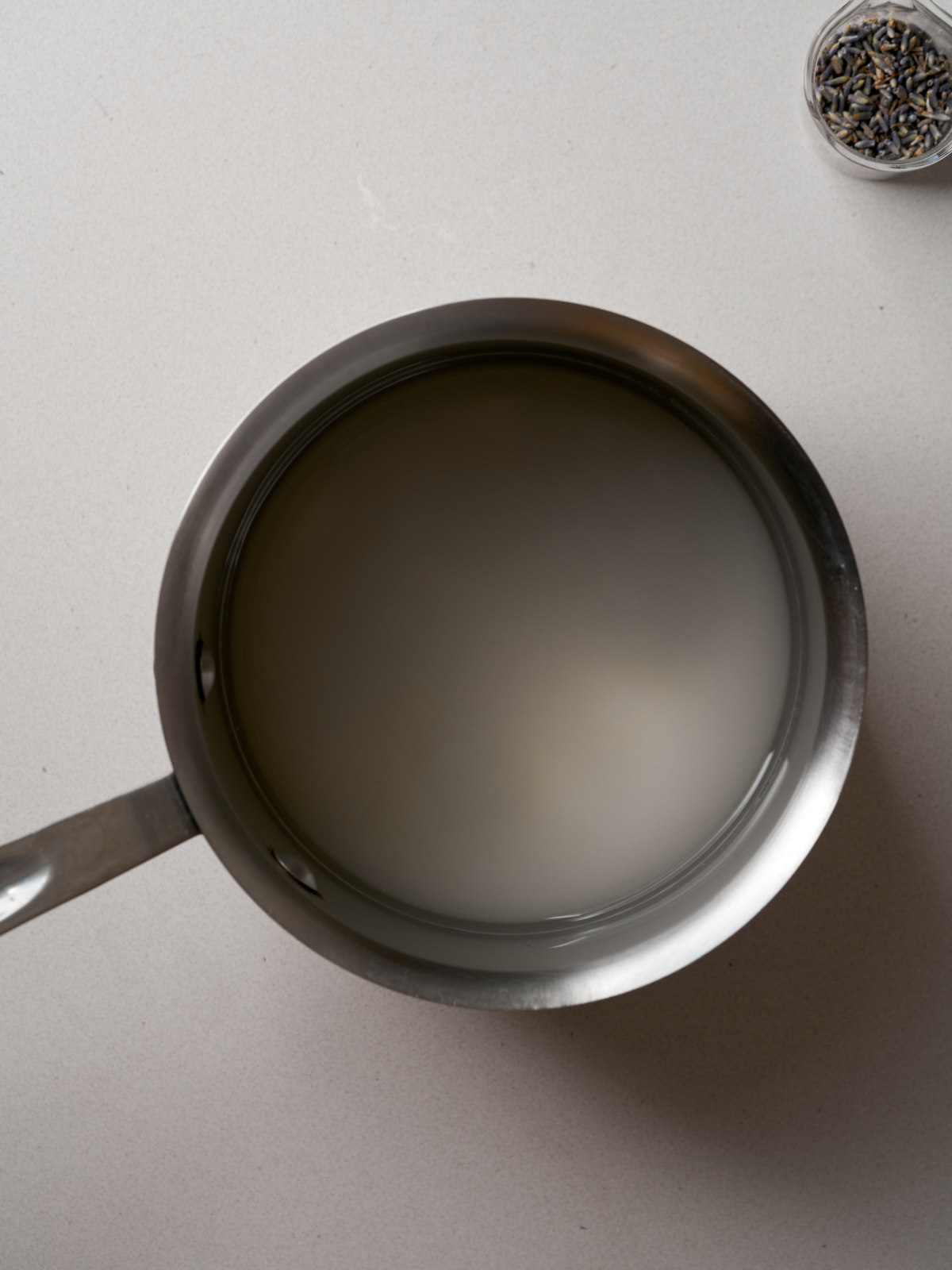 Sugar and water in a saucepan next to a small bowl of dried lavender flowers.