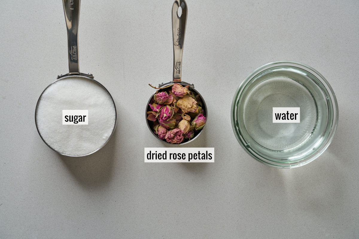 A measuring cup filled with sugar next to a measuring cup with dried roses next to a jar of water.