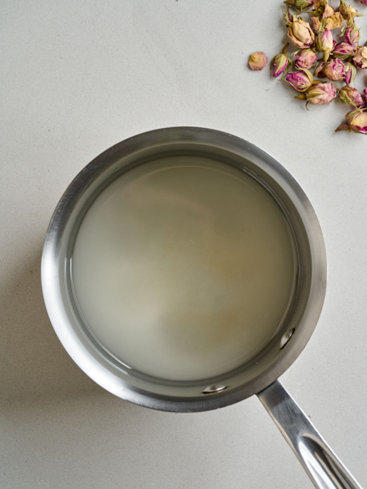 A saucepan with sugar and water next to dried rose petals.