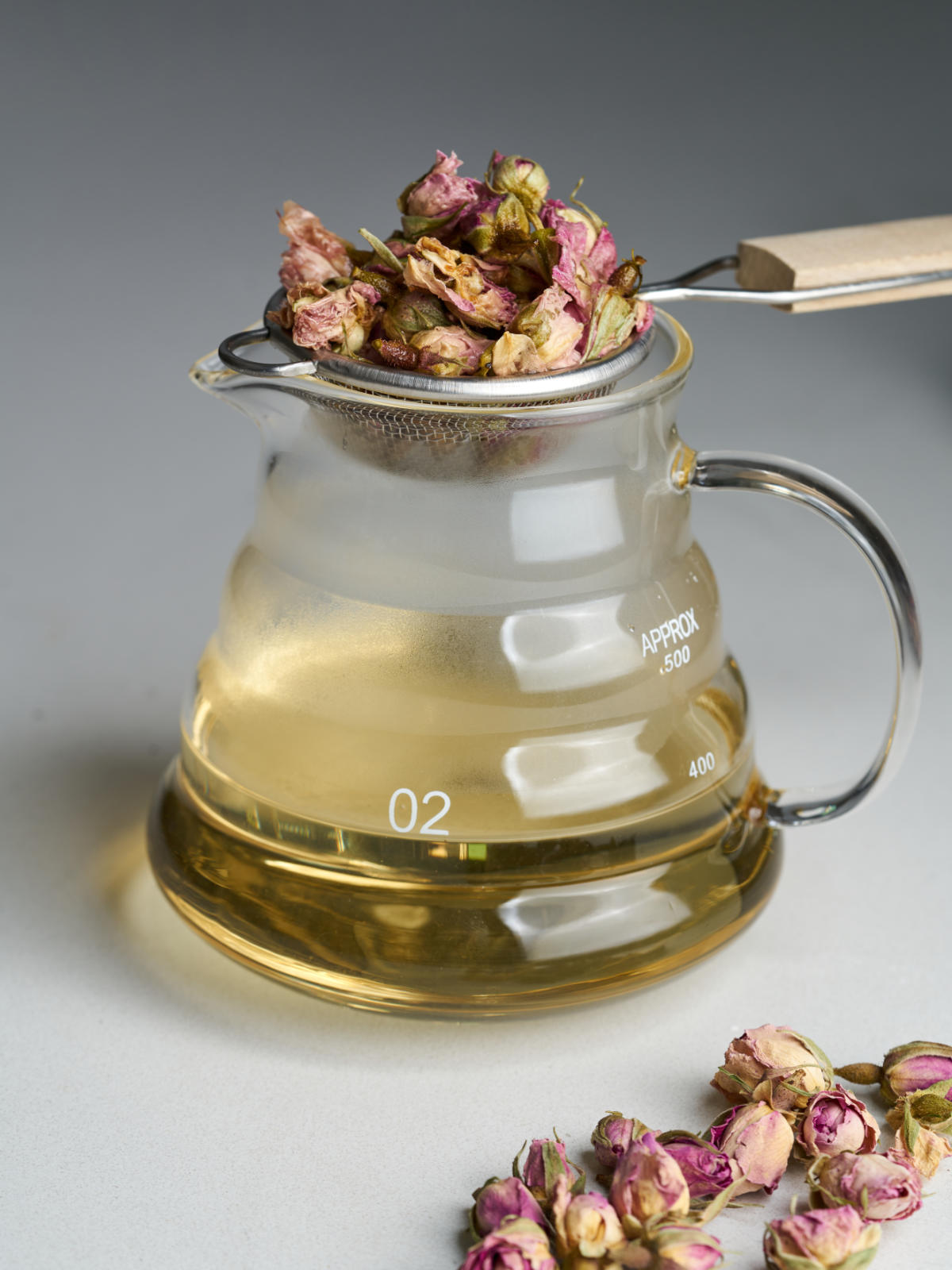 A glass pitcher with yellow simple syrup next to dried rose petals and with a strainer filled with dried roses on top of the pitcher.