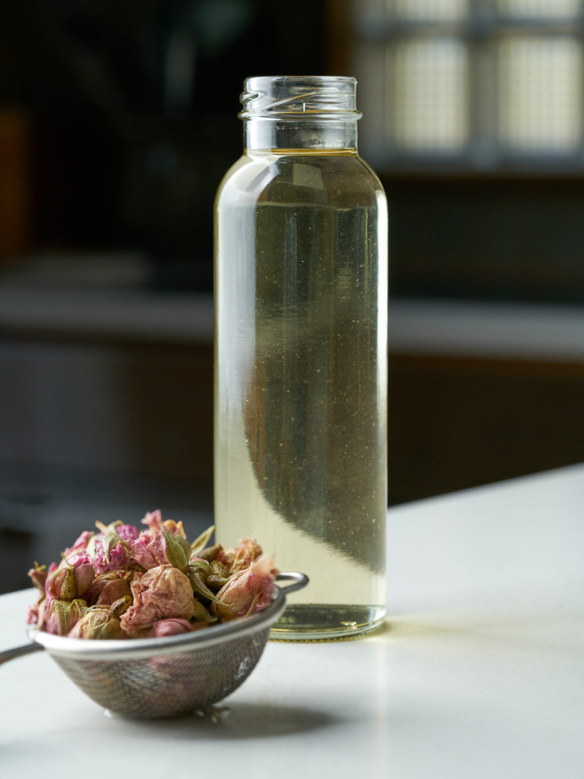 A bottle of yellow simple syrup next to a small strainer with dried roses.