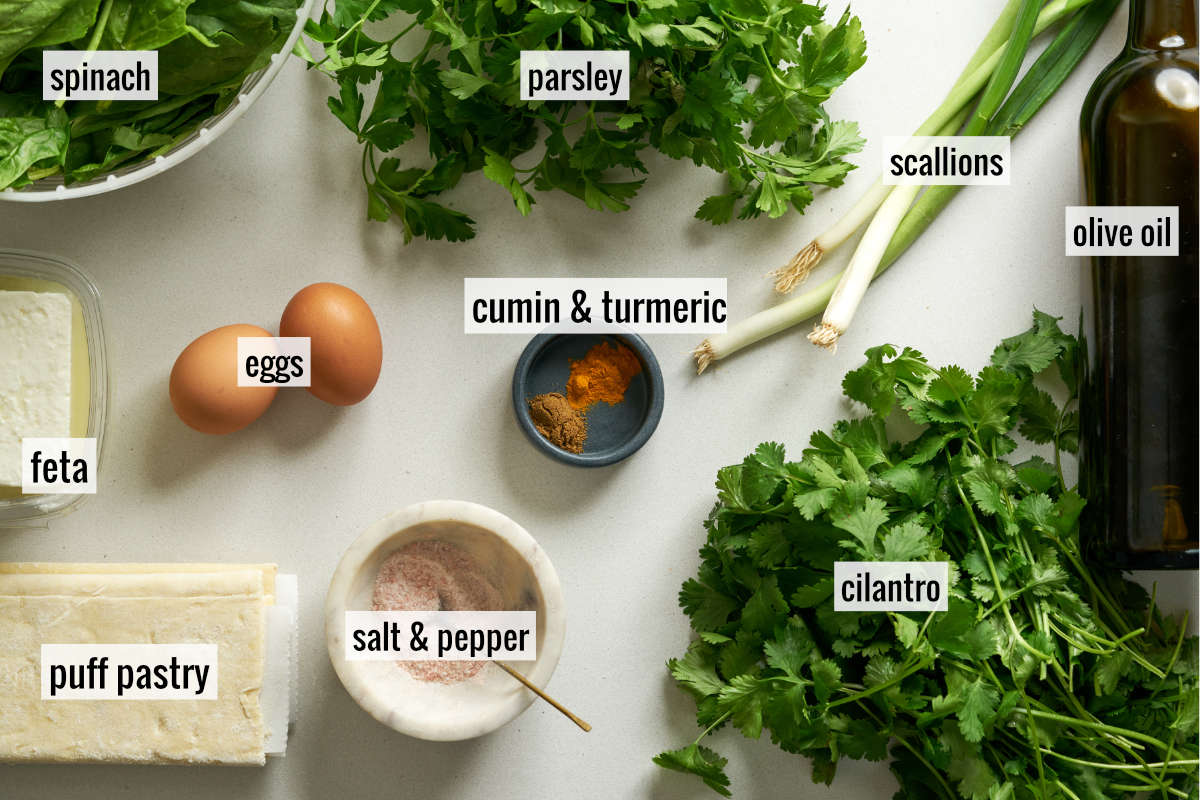 Herbs, feta, spices, eggs, and puff pastry on a countertop.