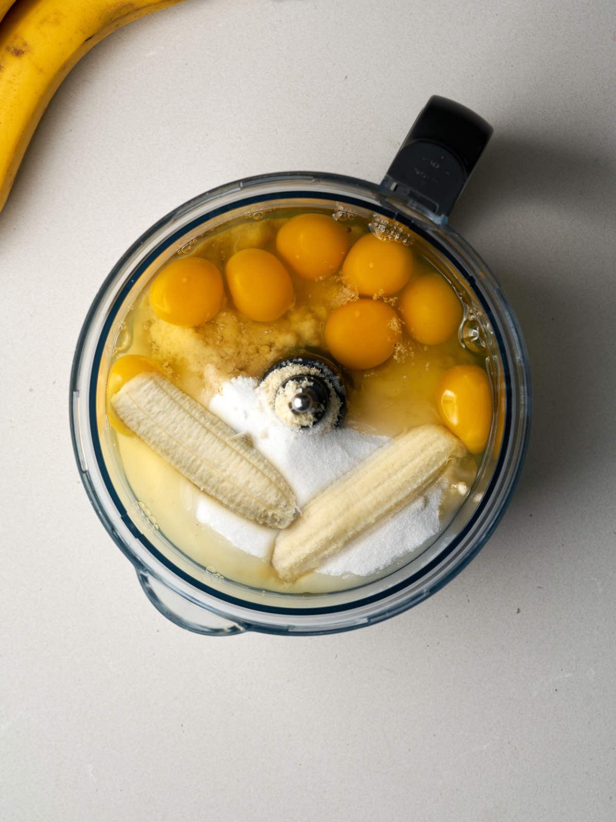 A mixture of eggs, sugar, flower, and bananas in a food processor with a black handle.