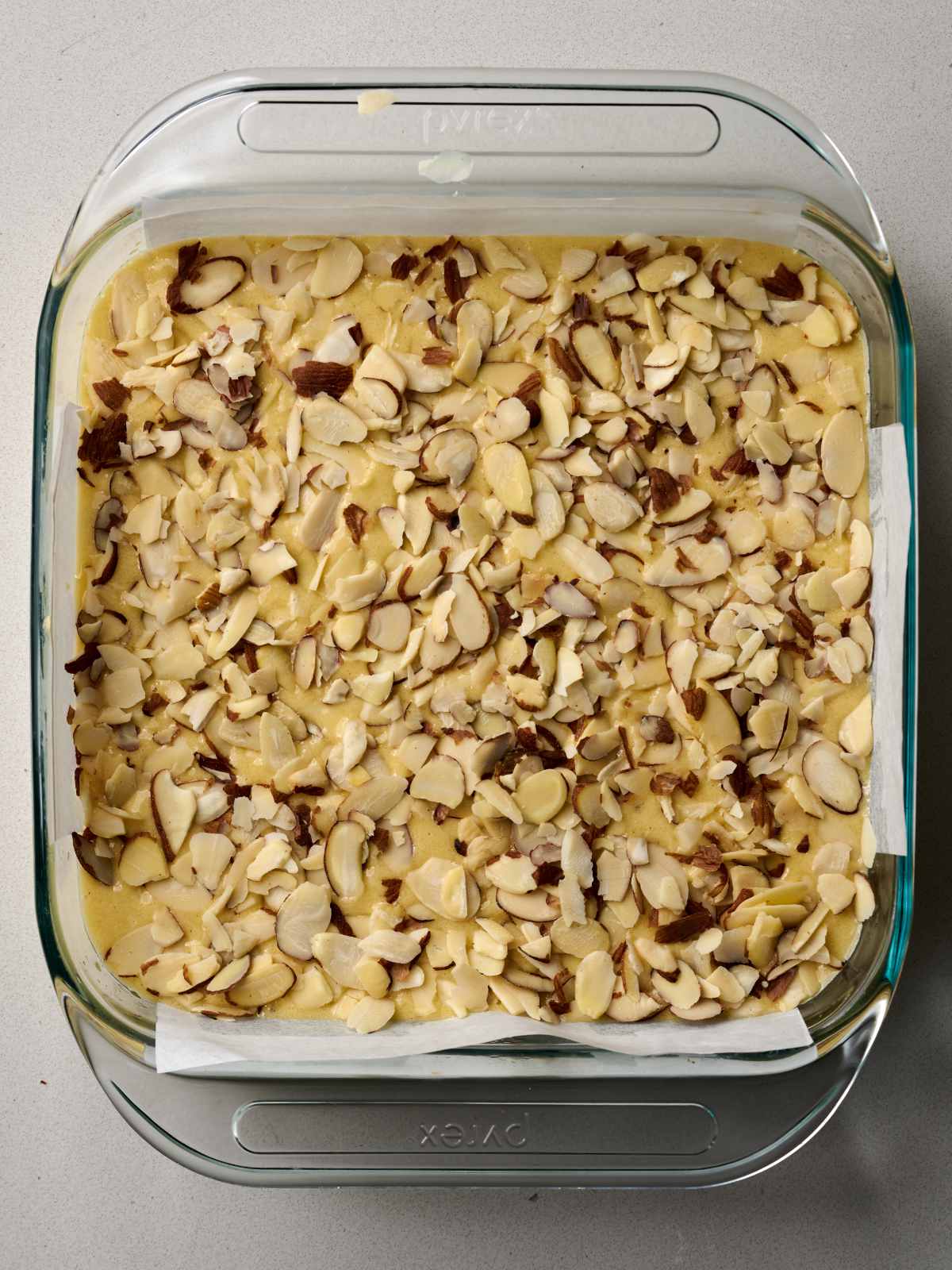 Raw batter in a glass baking dish lined with parchment paper covered in sliced almonds.