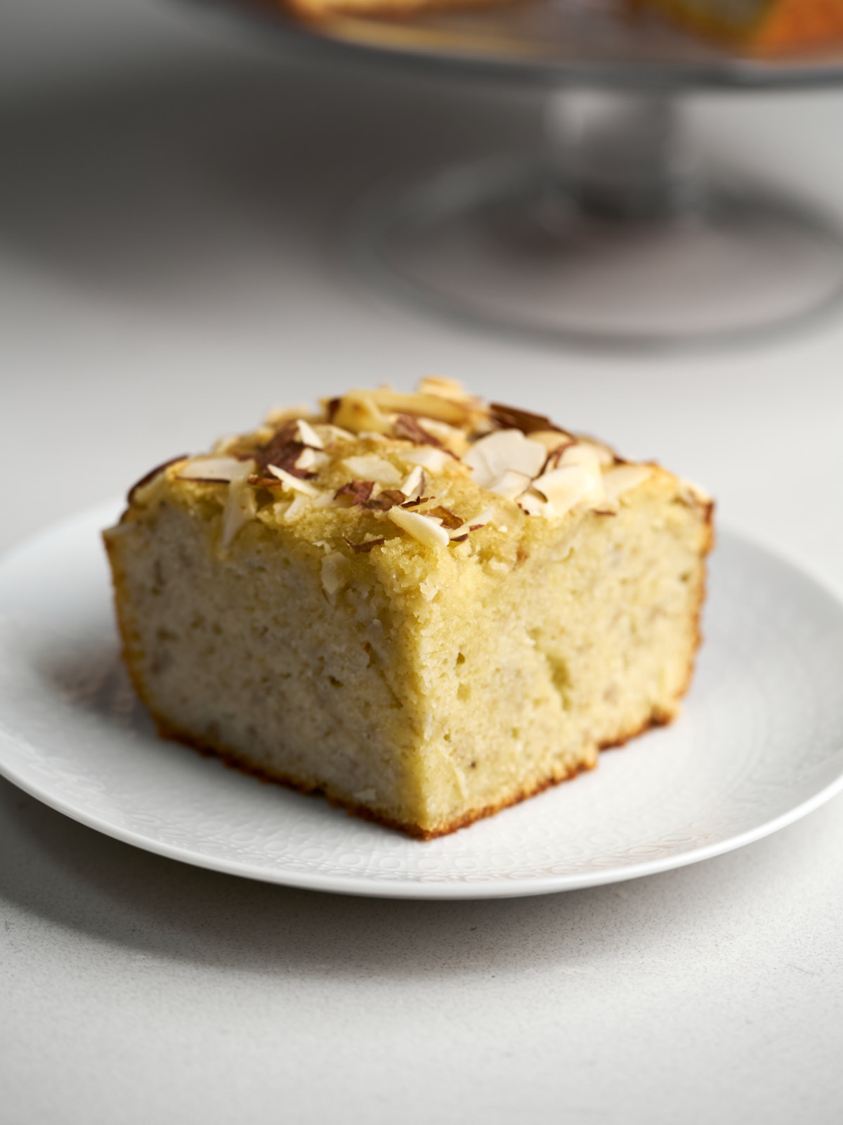 A square slice of cake with an almond topping on a white plate.