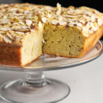 A beige square cake with an almond topping sitting on a glass cake display stand