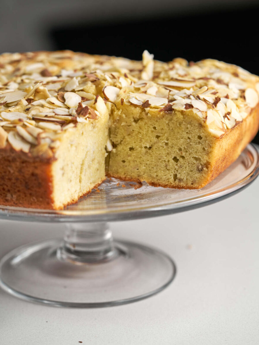 A beige square cake with an almond topping sitting on a glass cake display stand