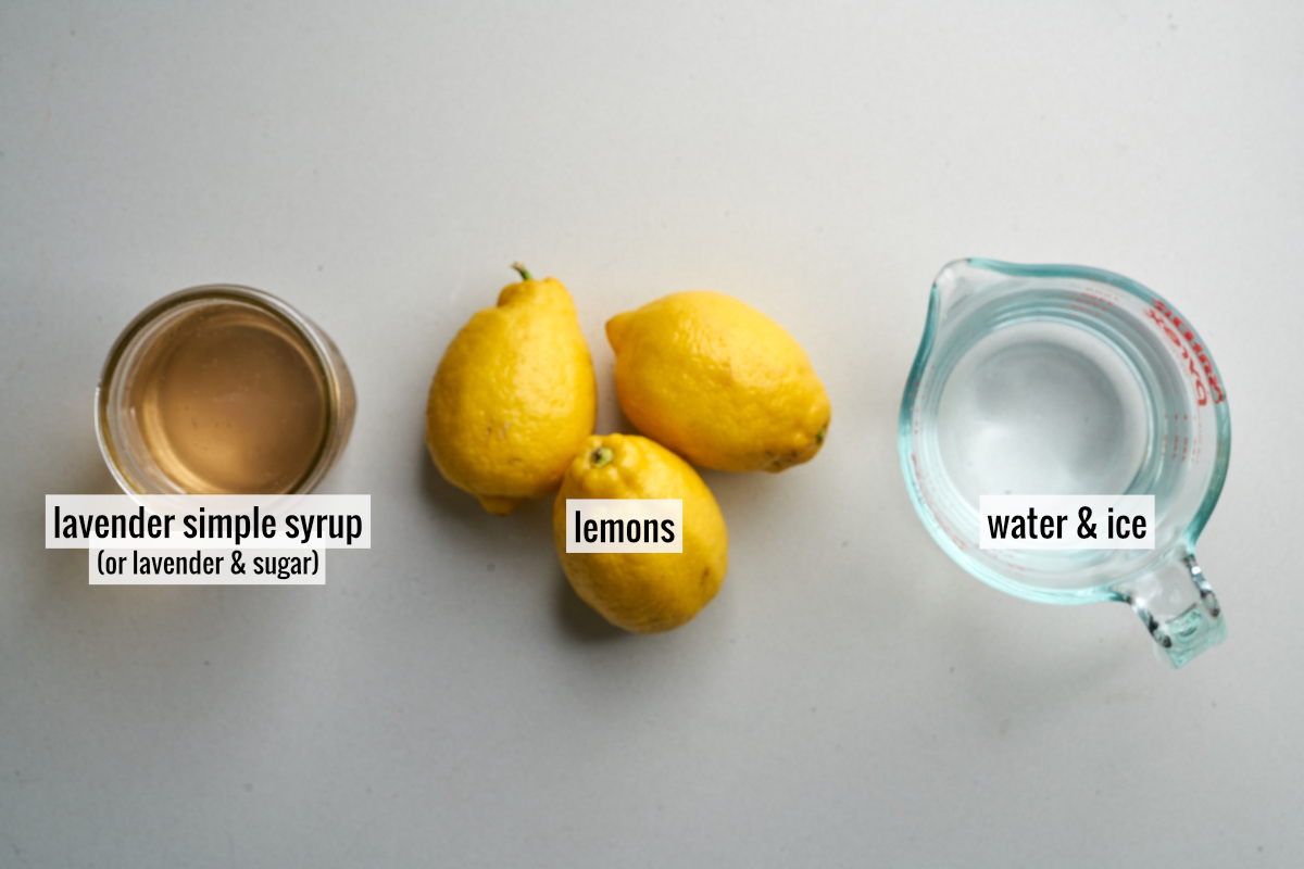 Three lemons next to a liquid measuring cup with water and a small jar of purple tinted simple syrup.