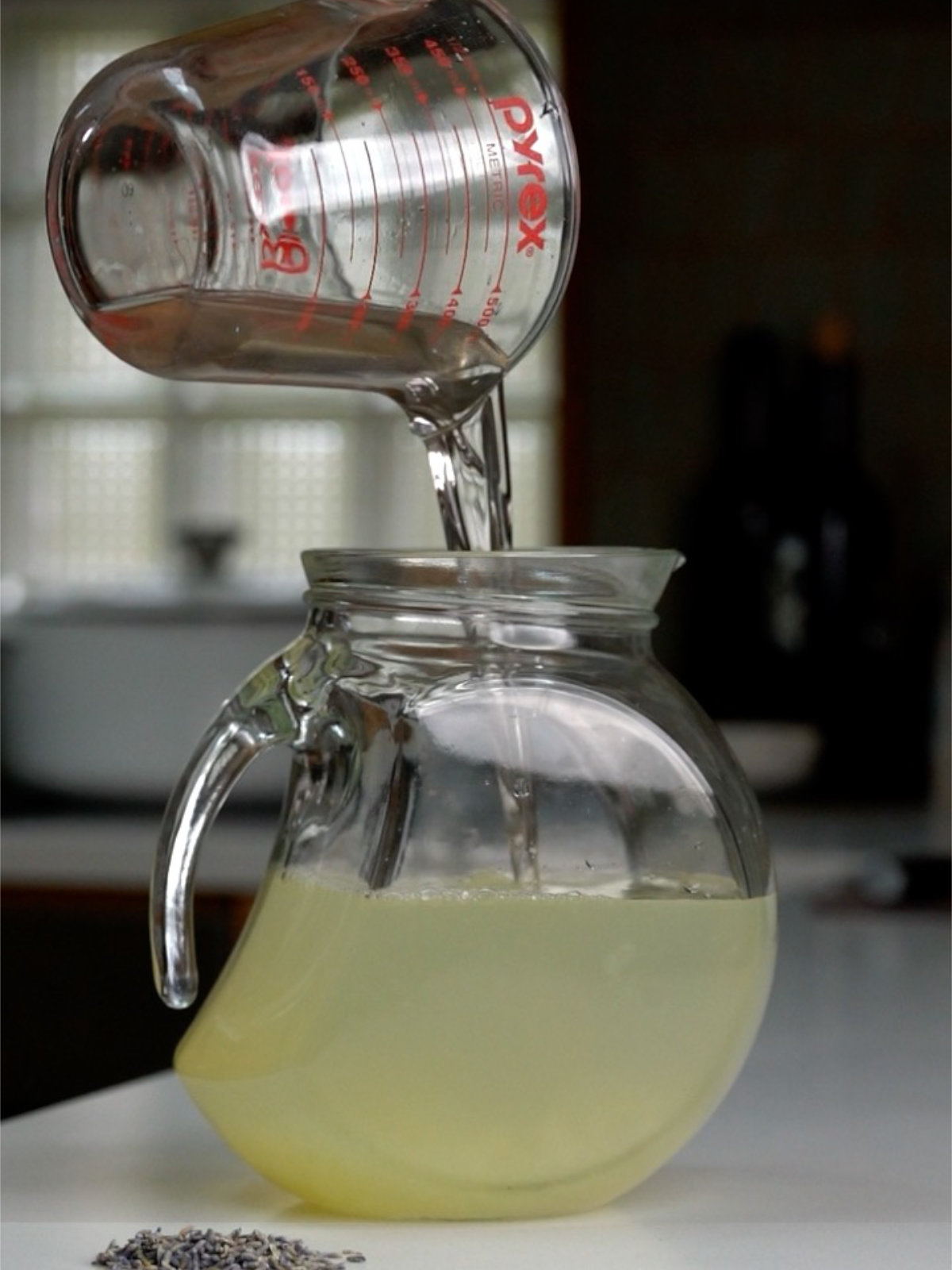 Pouring liquid into a glass pitcher from a liquid measuring cup on a countertop next to lavender flowers.