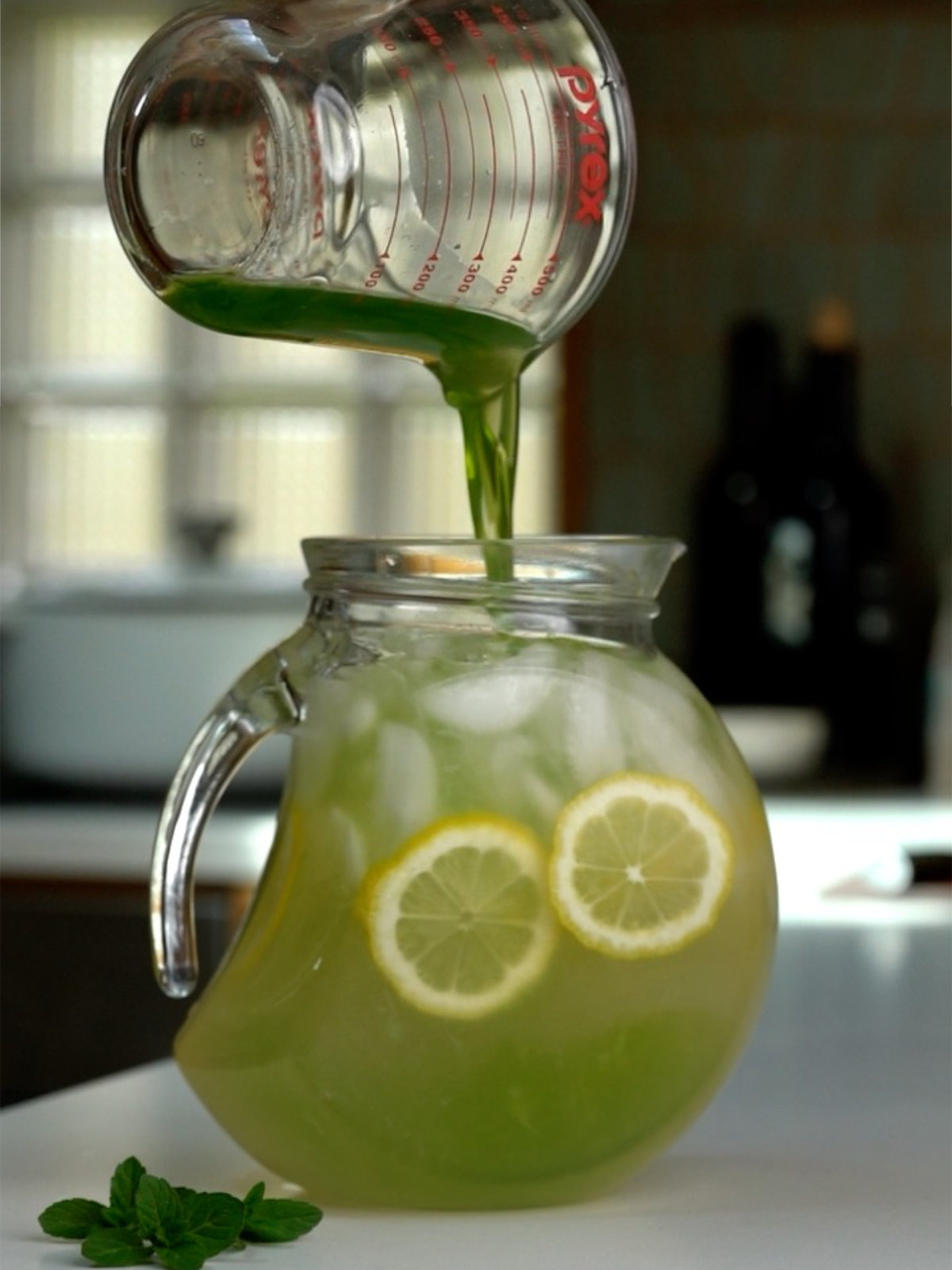 Pouring green liquid into a glass pitcher from a liquid measuring cup on a countertop next to mint leaves.