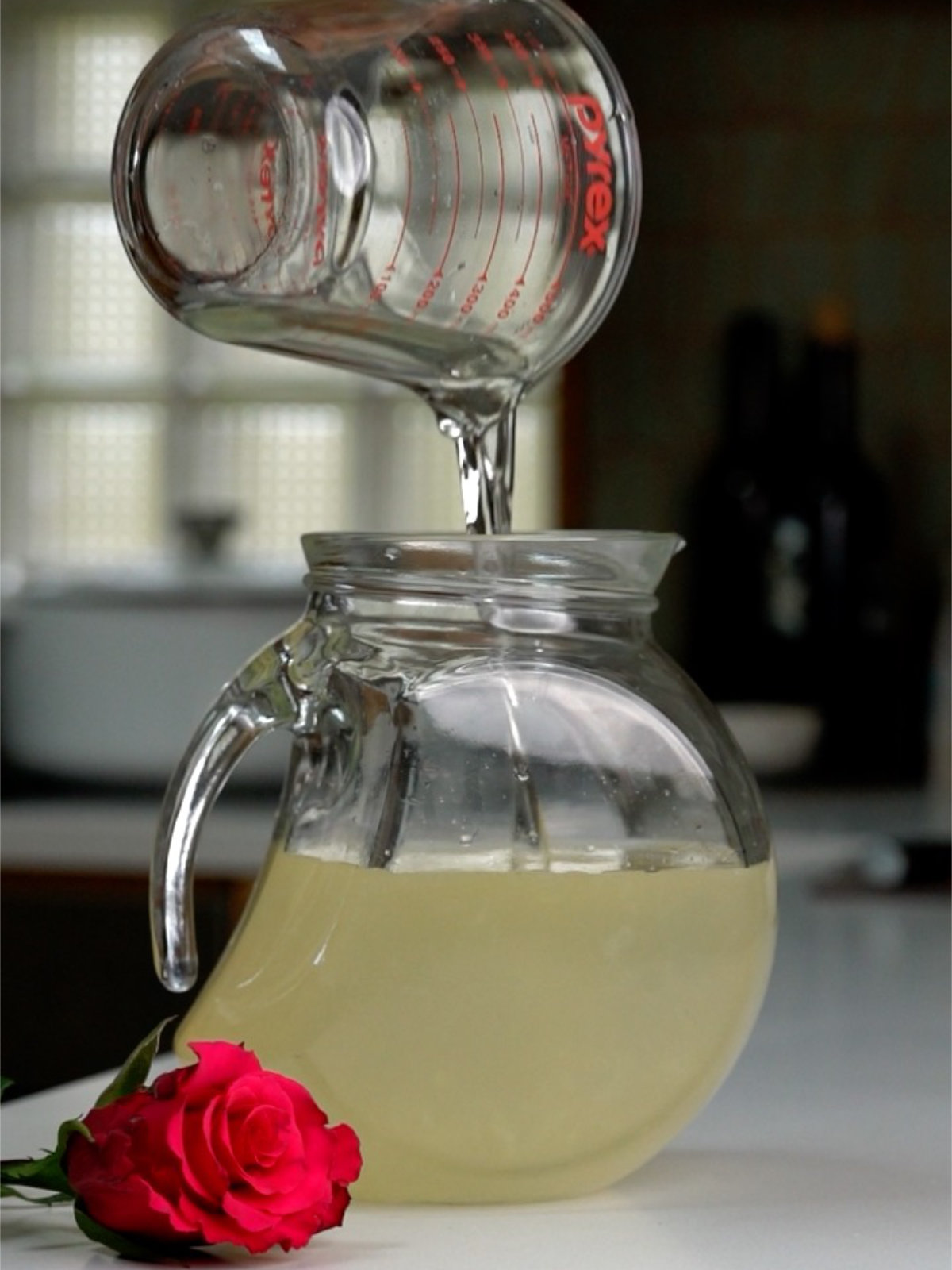 Pouring liquid into a glass pitcher from a liquid measuring cup on a countertop next to a hot pink rose.