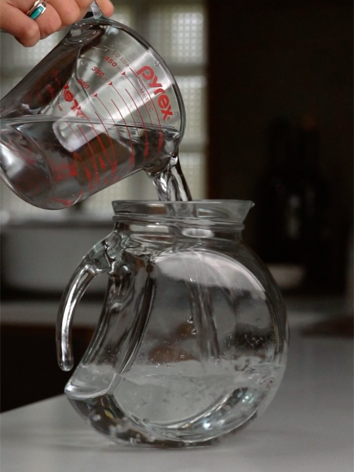 Pouring water into a glass pitcher from a liquid measuring cup on a countertop.