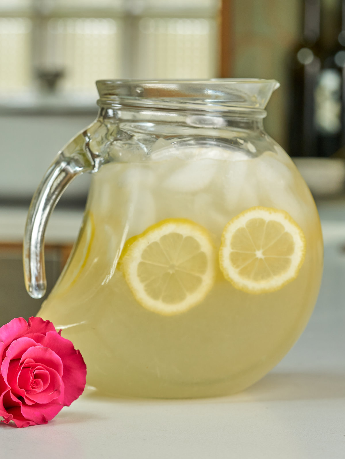 Pitcher with lemonade, lemon slices, and ice next to a hot pink rose.