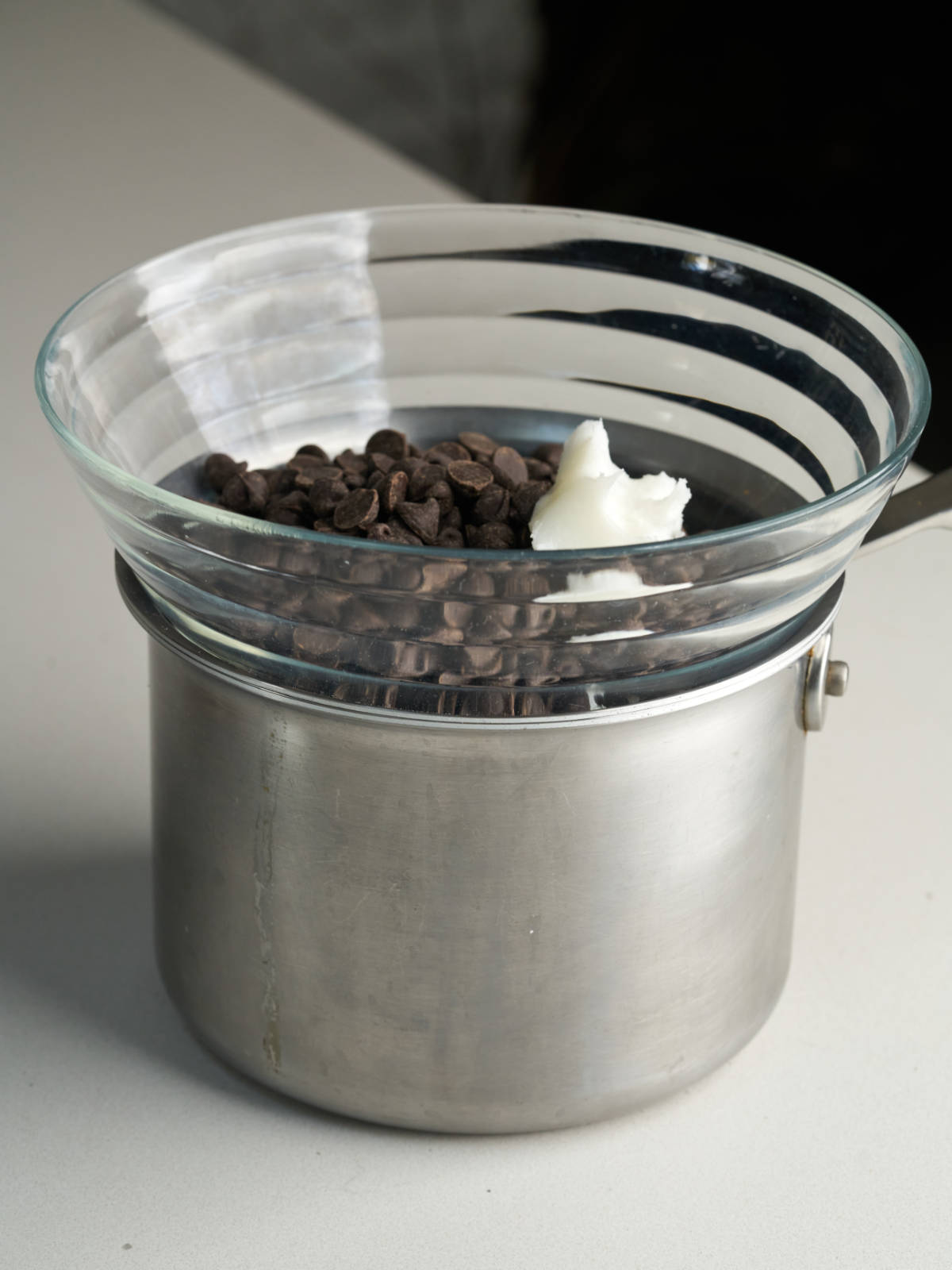 A glass bowl with chocolate chips and coconut oil sitting on top of a stainless steel saucepan.